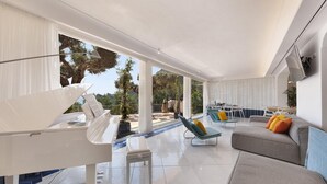 Ein modernes Wohnzimmer mit einem Klavier, einem grauen Sofa mit bunten Kissen, einem Essbereich mit weißen Stühlen und Blick auf Bäume und Berge durch große Fenster.