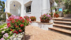 Una villa blanca con un muro de piedra, un patio con patrón de espiga y flores en macetas.