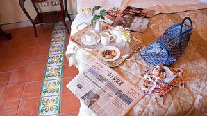 Un plateau de petit-déjeuner bien organisé, avec des tasses, une théière et une assiette de nourriture, est posé sur un lit recouvert d’une courtepointe à motifs. Un journal est étalé sur le lit, et un sac bleu à motifs est visible.
