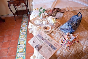 A neatly arranged breakfast tray with cups, a teapot, and a plate of food on a bed with a patterned bedspread. A newspaper is spread out on the bed, and a blue patterned bag is visible.