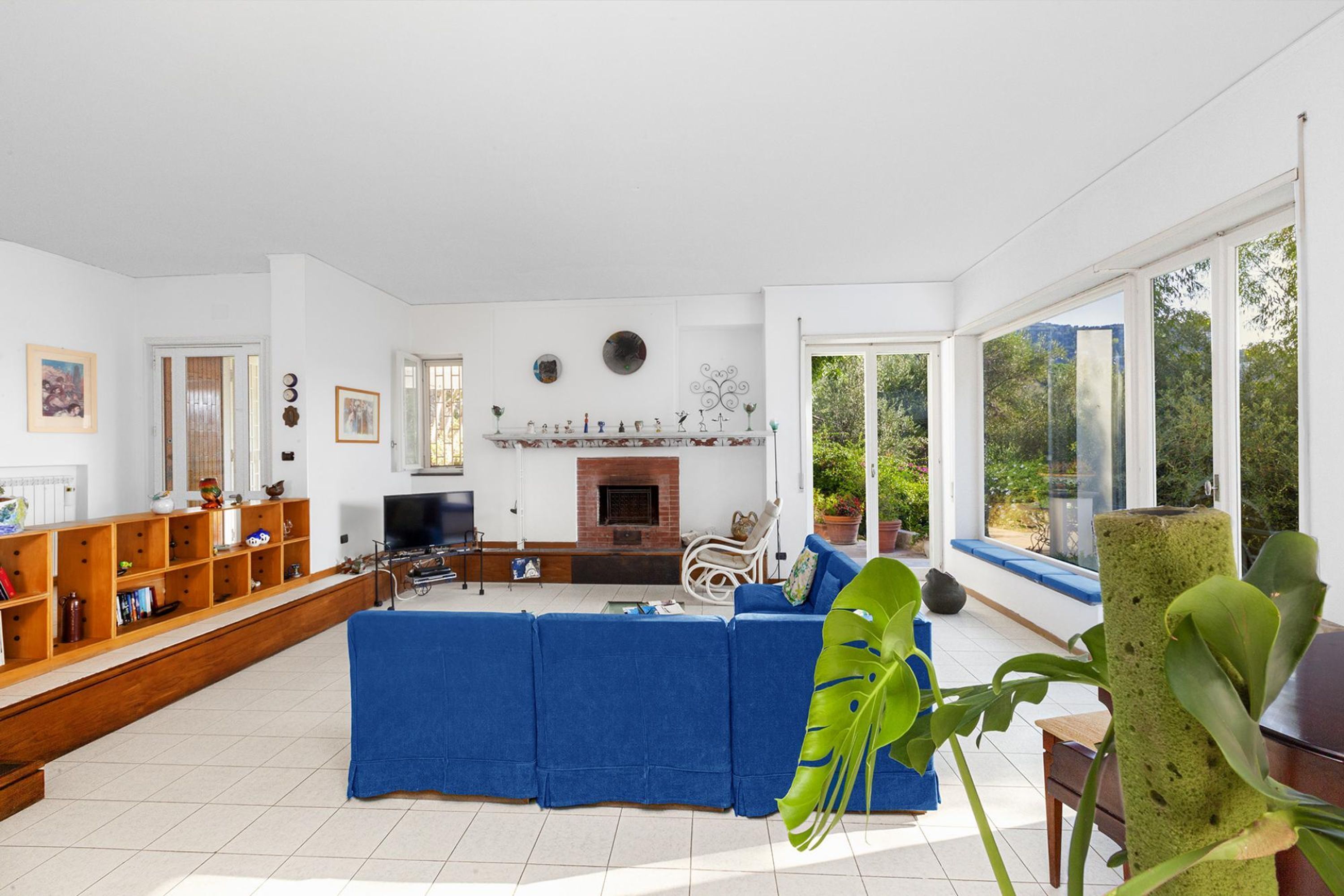 Uma sala de estar ampla com sofá azul, lareira, televisão e uma grande janela com vista para o verde.