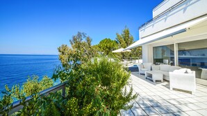 Een modern terras buiten met witte meubels, een glazen balustrade en uitzicht op zee.