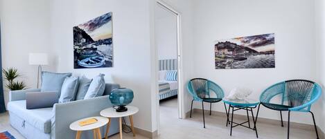 A modern living room with a sofa, two side tables, a lamp, and two chairs. There are two paintings on the walls and a view into another room.