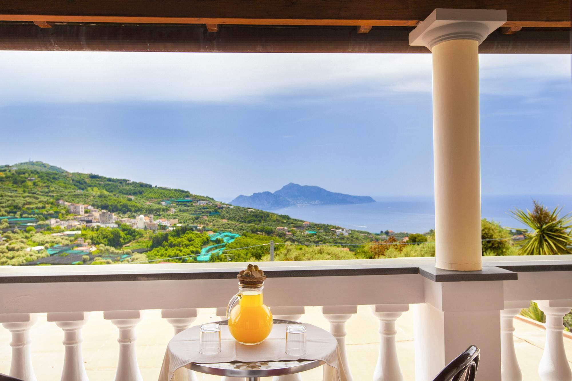 A balcony with a table, a bottle of orange juice, and a view of a coastal landscape with mountains and greenery.