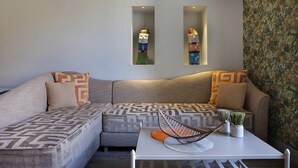 Ein modernes Wohnzimmer mit einem grauen Ecksofa, gemusterten Kissen, einem weißen Couchtisch und einem Metallstuhl sowie zwei Wandregalen mit Dekorationsobjekten.