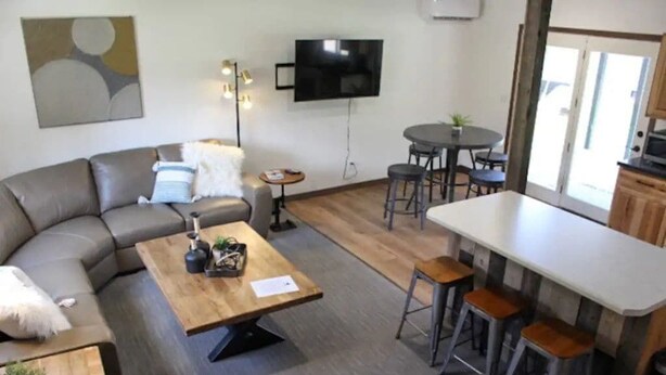 A modern living room with a sofa, coffee table, and bar stools. A flat-screen TV is mounted on the wall, and there is a small round table with chairs.