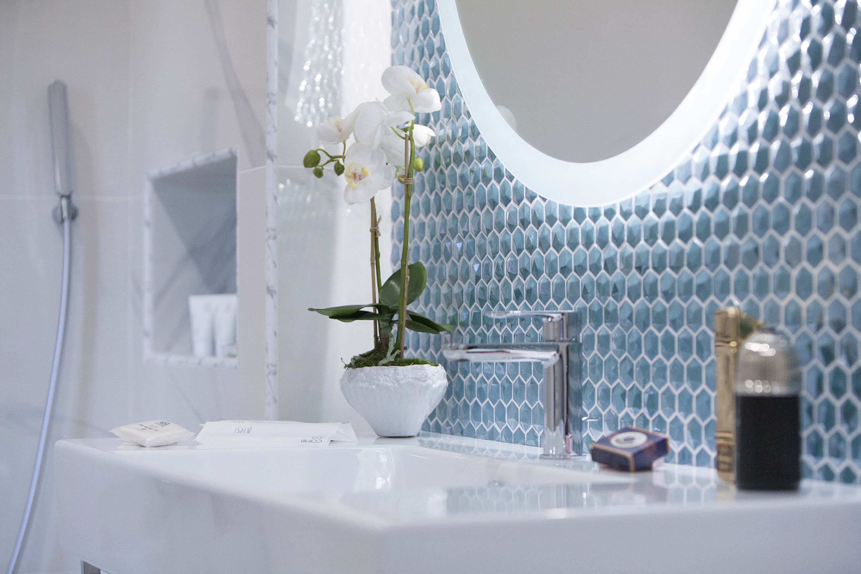 A bathroom with a white sink, a white vase with an orchid, a glass bottle, and a mirror with a blue hexagonal tile backsplash.
