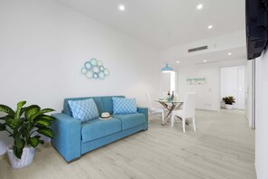 Uma sala de estar moderna com um sofá azul, almofadas estampadas em padrão geométrico, uma mesa de centro de vidro e uma área de jantar com cadeiras brancas.