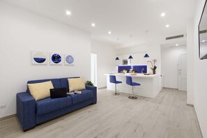 Sala moderna com sofá azul, área de cozinha com bancada branca e bar com cadeiras azuis.