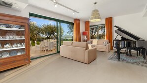 A spacious living room with a piano, a sofa, and a dining area with a glass door leading to a garden. - Villa Elia in Massa Lubrense (Massa Lubrense)
