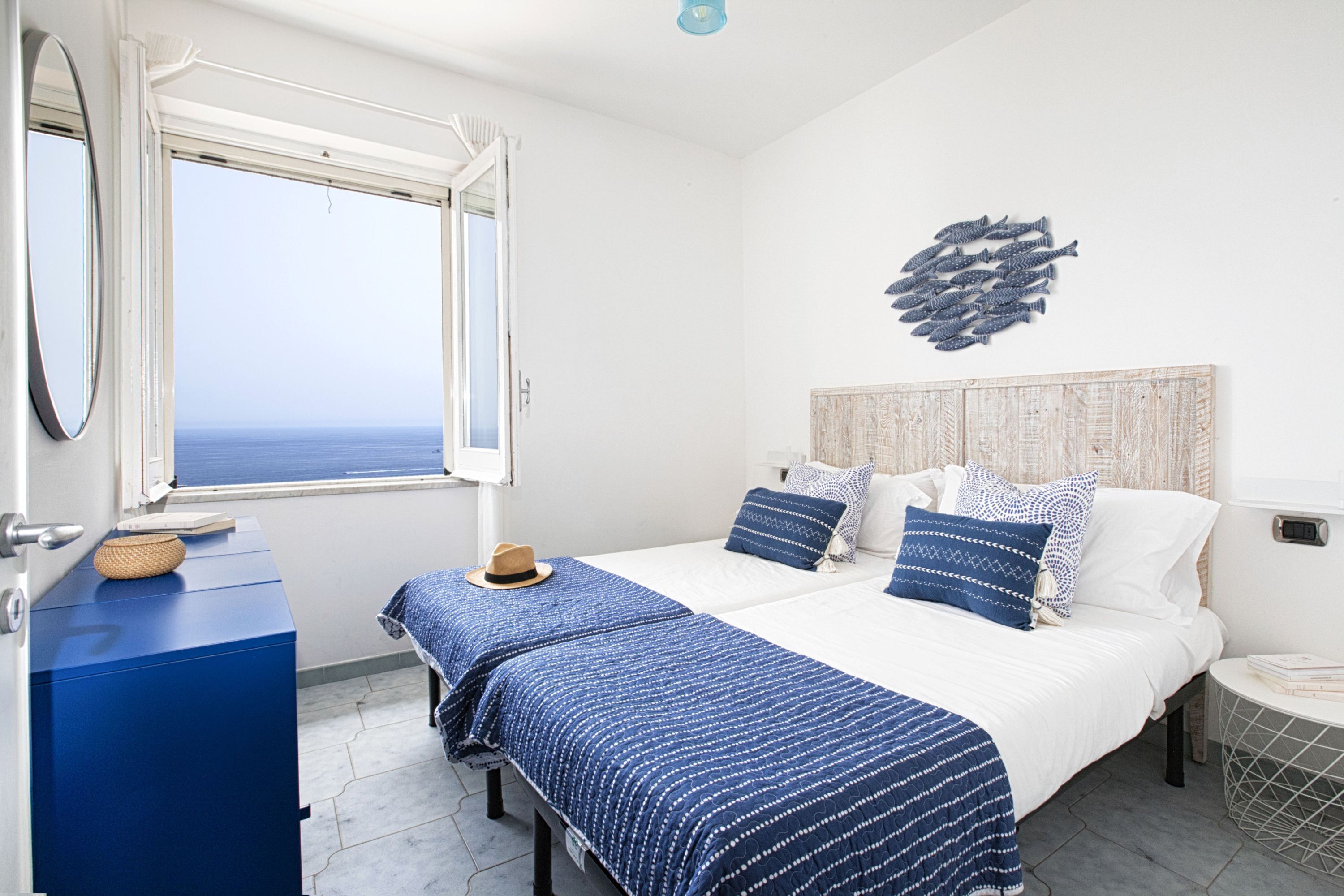 Un dormitorio con un ventanal grande, una cama con ropa de cama azul, una mesita de noche y una obra de arte en la pared.