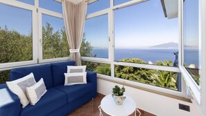 Ein blaues Sofa mit weißen Kissen, ein kleiner weißer Tisch mit einer Pflanze und große Fenster mit Blick auf das Meer und die grüne Landschaft.