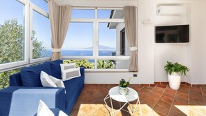 Ein Zimmer mit einem blauen Sofa, einem kleinen weißen Tisch, einer Topfpflanze, einem an der Wand montierten Fernseher und einem großen Fenster mit Vorhängen.
