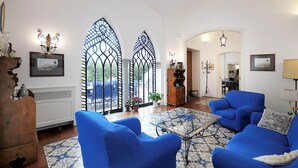 A living room with blue armchairs, a glass coffee table, and a patterned rug. There are two stained glass windows, a wooden cabinet, and a door leading to another room. - Villa Angelo in Sant Agnello (Sant'Agnello)