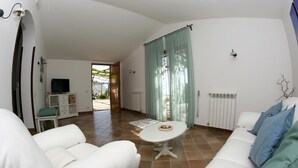 A spacious living area with a white sofa, a small round table, and a TV on a white cabinet. There is a door leading to an outdoor area with a view of mountains. - Villa Raffaella in Sant Agata sui Due Golfi (Massa Lubrense)