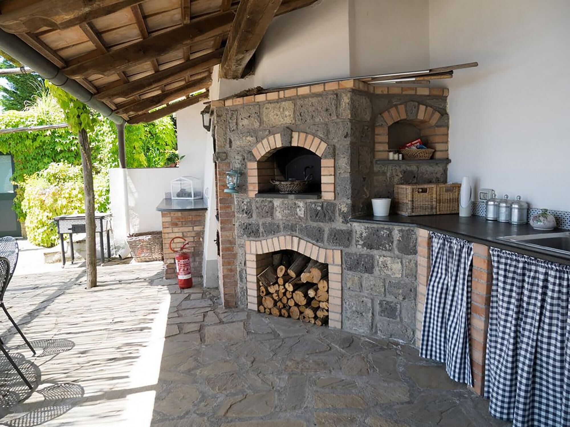 Forno de pizza de pedra com arco de tijolos, um extintor de incêndio e uma mesa com cadeiras sob uma pérgola de madeira.