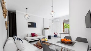A modern living room with a white sofa, dining table, and chairs. There are two pendant lights, a framed picture on the wall, and a window with curtains. - Appartamento Sorrento Suite de Charme 1 (Sorrento)