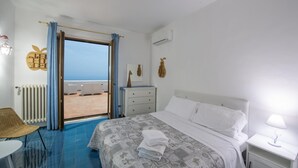 A hotel room with a bed, a nightstand, a chair, a table, and a view of the sea through an open door. - Villa Isabel in Praiano (Praiano)
