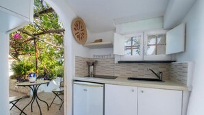 A compact kitchen with white cabinets, a sink, and a small refrigerator. There is a dining table with chairs outside, and a decorative wall piece above the sink. - Villa Isabel in Praiano (Praiano)
