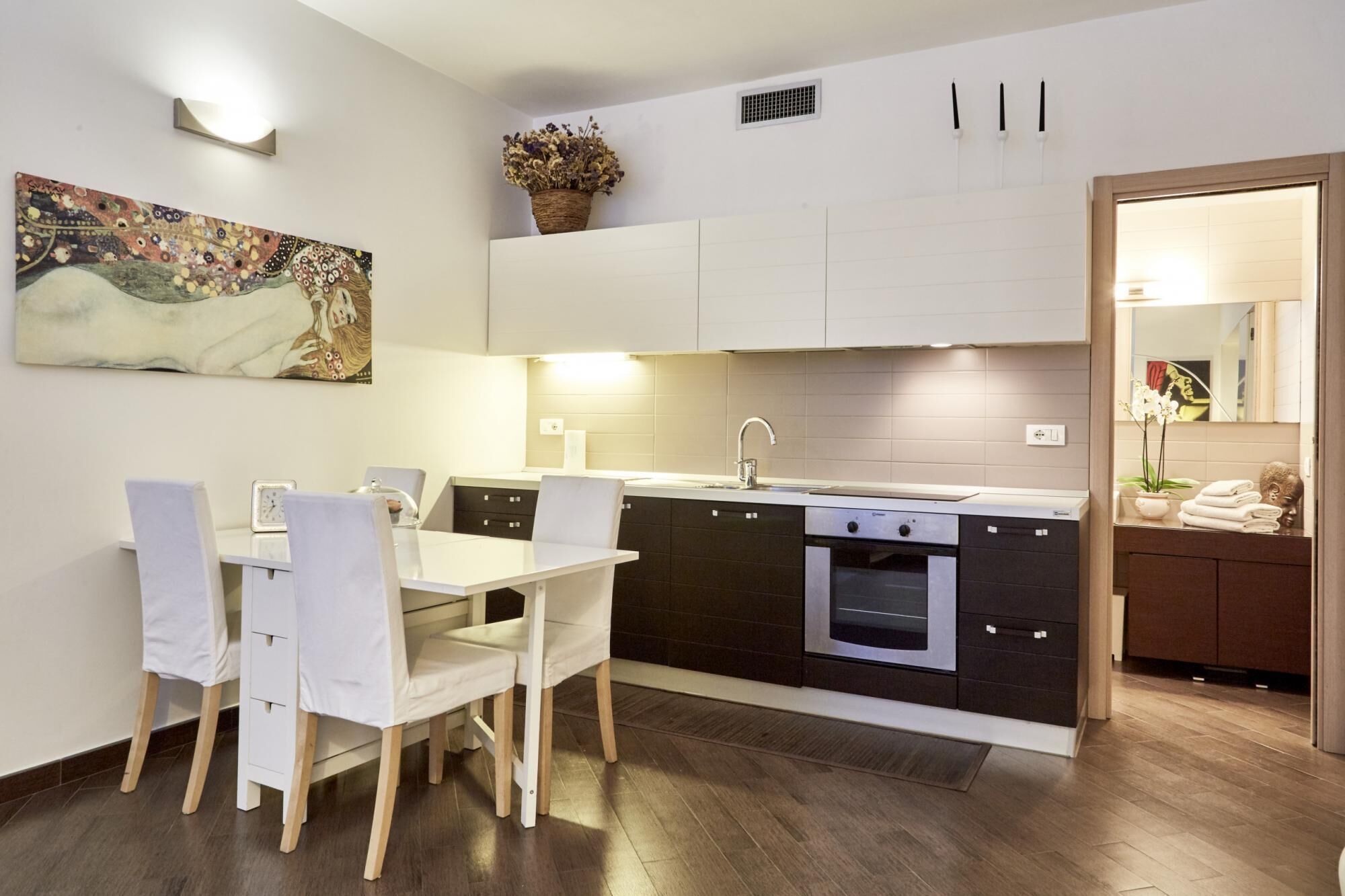 A modern kitchen with a dining area, featuring a white table, chairs, and a painting on the wall.