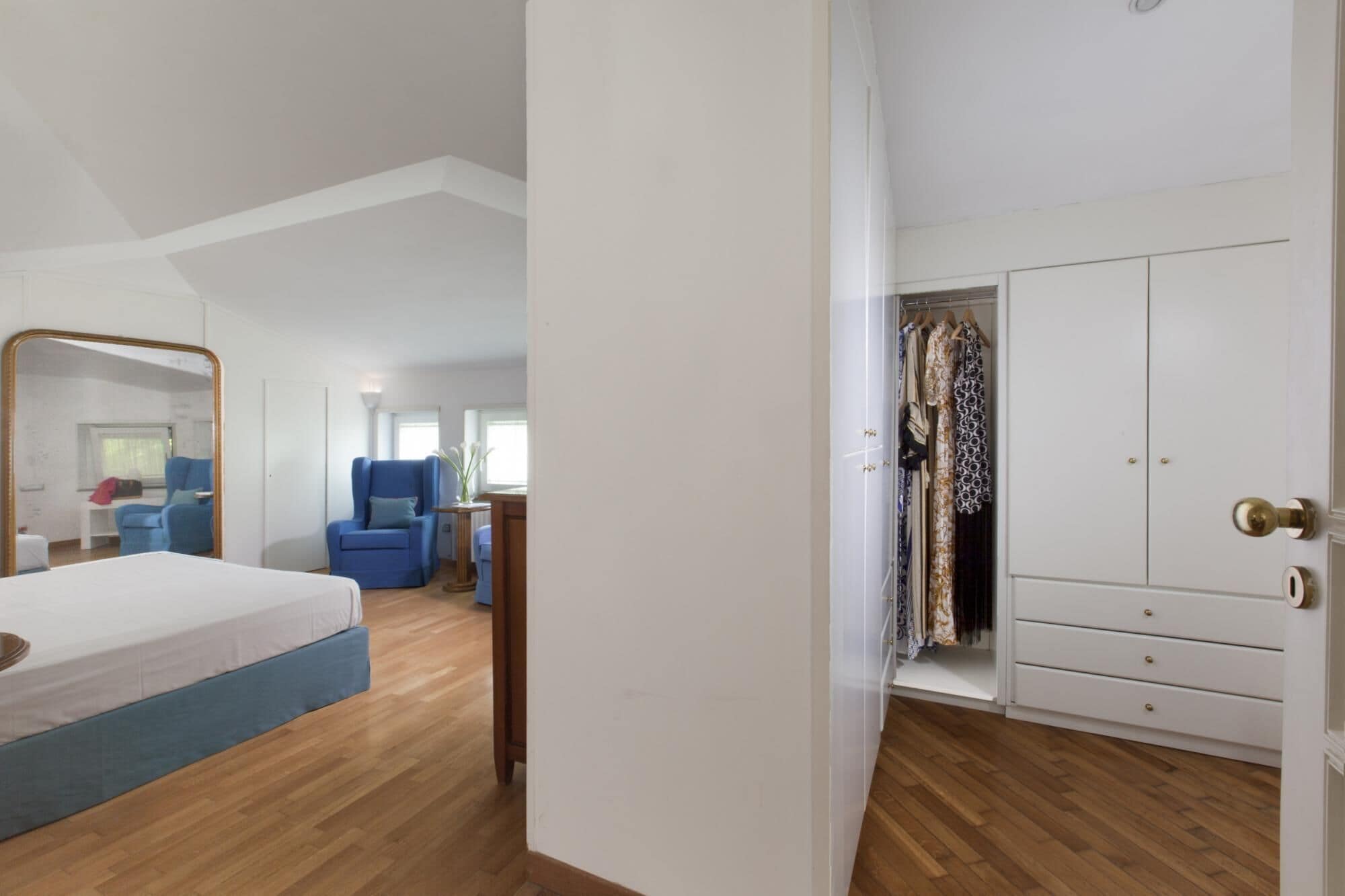 A bedroom with a bed, a blue armchair, a small table with a vase, and a wardrobe with hanging clothes.