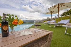 A wooden table with a bottle, glasses, and flowers by a poolside with lounge chairs and umbrellas. - Ville Il Paradiso - Villa Bacci (Greve in Chianti)