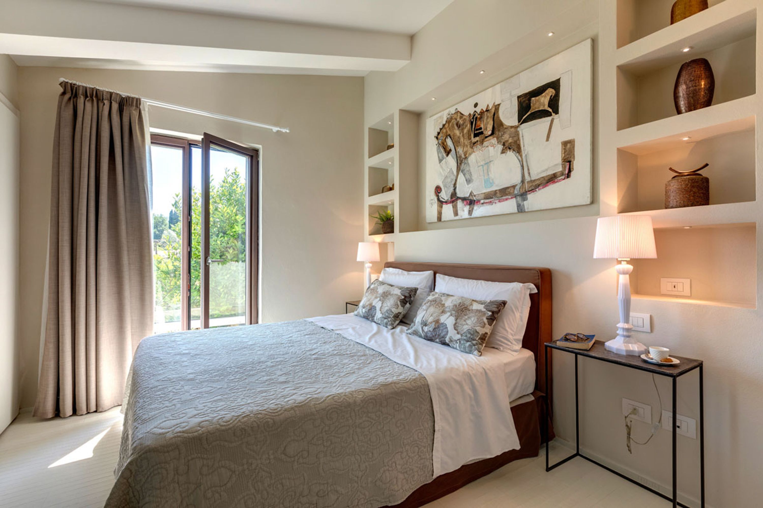 Une chambre avec un grand lit, une fenêtre avec des rideaux, une table de chevet avec une lampe et une étagère murale décorative.