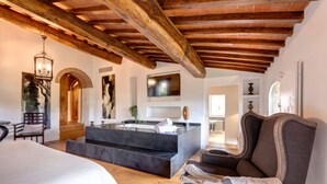A spacious room with a large bed, a seating area with a sofa and armchairs, a built-in bathtub, wooden beams on the ceiling, and a flat-screen TV mounted on the wall. - Ville Il Paradiso in Panzano (Greve in Chianti)