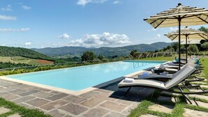 A swimming pool with lounge chairs and umbrellas, surrounded by stone tiles and greenery, with a view of distant hills. - Ville Il Paradiso in Panzano (Greve in Chianti)
