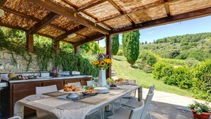 An outdoor dining area with a stone wall, a wooden pergola, wicker chairs, and a view of a green landscape. - Ville Il Paradiso in Panzano (Greve in Chianti)
