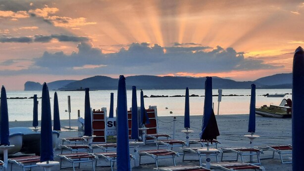 A beach with rows of sun loungers and umbrellas, a lifeguard stand, and a calm sea with distant mountains. - Casa Alice 1 Bedrooms Apartment in Alghero (Alghero)