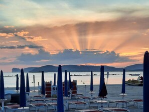 Une plage bordée de rangées de chaises longues et de parasols, un poste de surveillance pour les sauveteurs, et une mer calme avec des montagnes qui se dessinent à l’horizon.