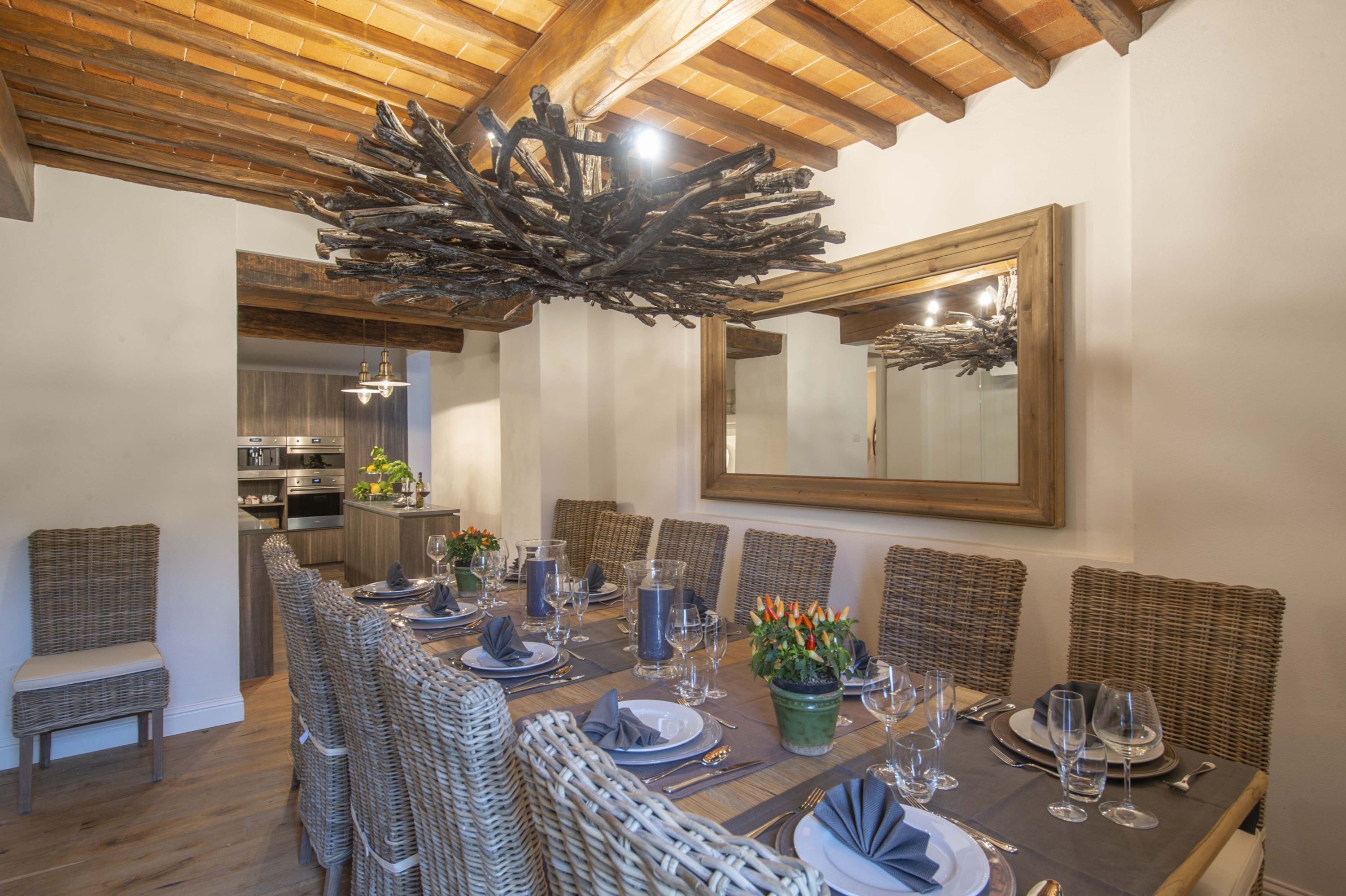 A dining area with a long table set for a meal, surrounded by wicker chairs, a wooden ceiling, and a large mirror on the wall.