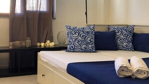 A neatly made bed with blue and white patterned pillows, a bedside table with decorative items, and a window with curtains. - Casa Ilaria 2 Bedrooms Apartment in Alghero (Alghero)