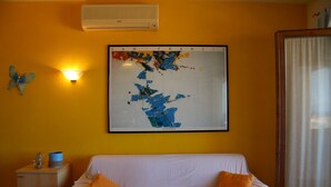 Une pièce avec un canapé blanc, deux coussins orange, un tableau abstrait encadré, un climatiseur fixé au mur et une petite table sur laquelle se trouve un récipient bleu.