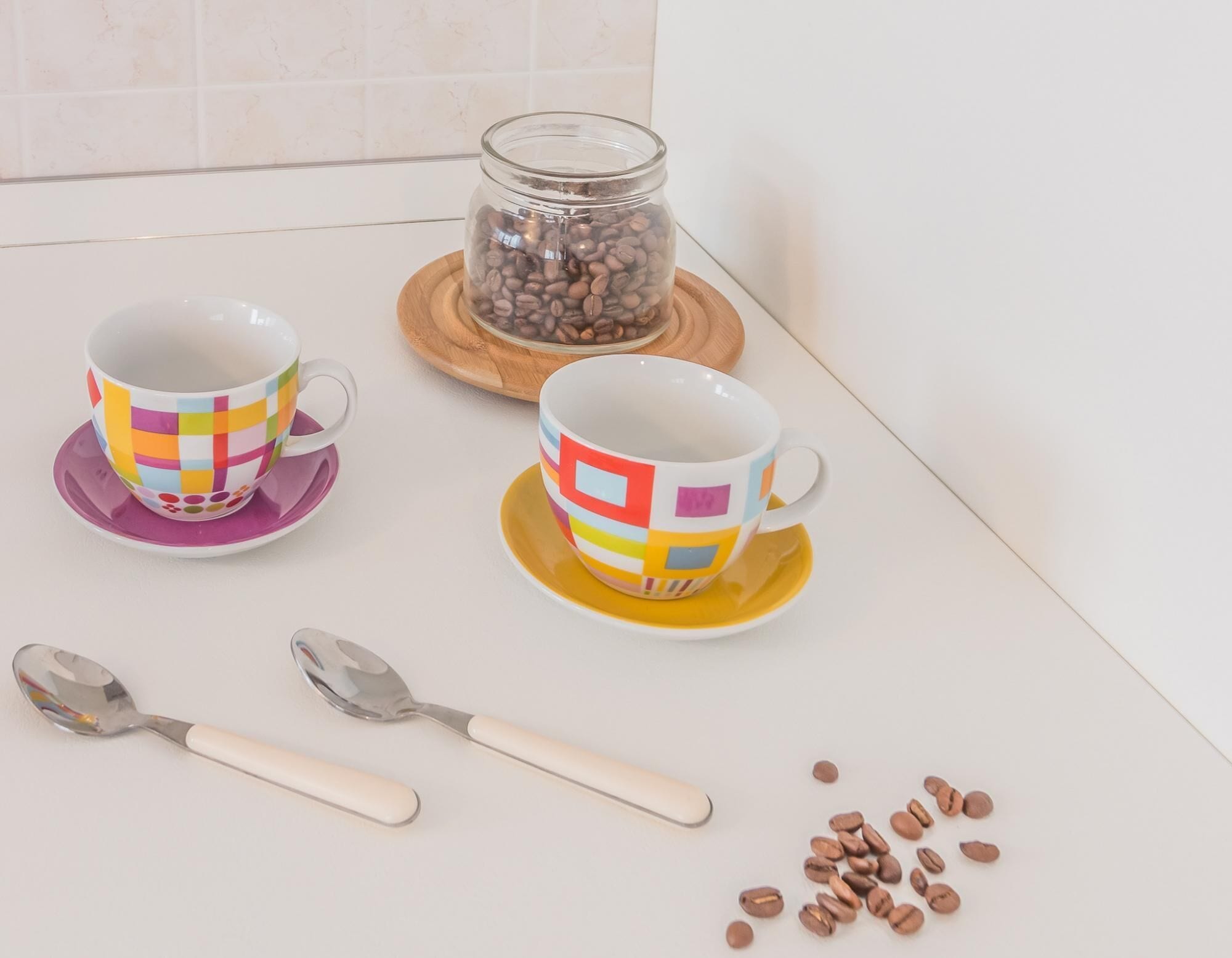Two colorful coffee cups on a white table with a spoon and scattered coffee beans.