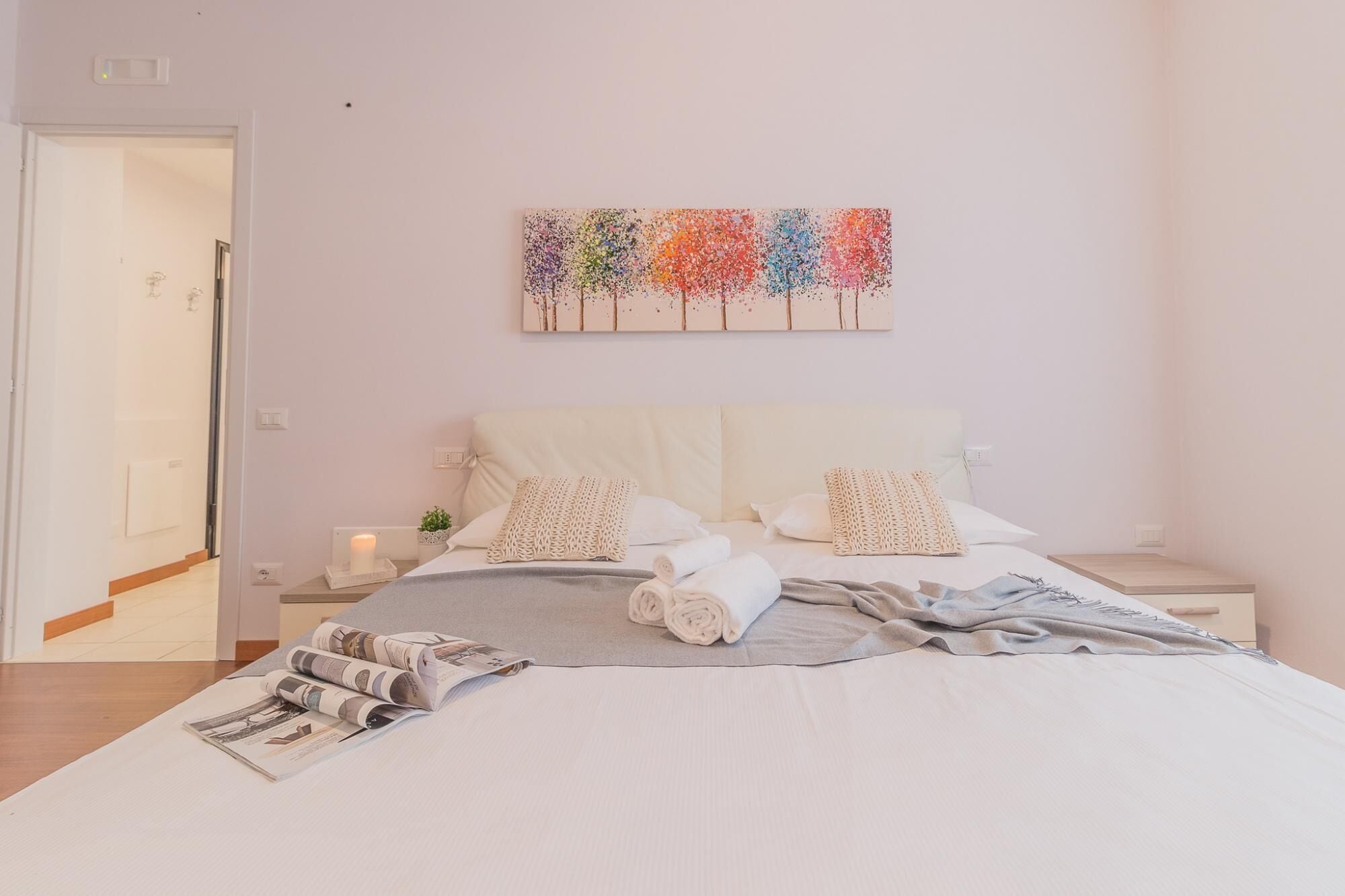A neatly made bed with white linens and a gray blanket, a bedside table with a candle, and a wall art piece above the bed.
