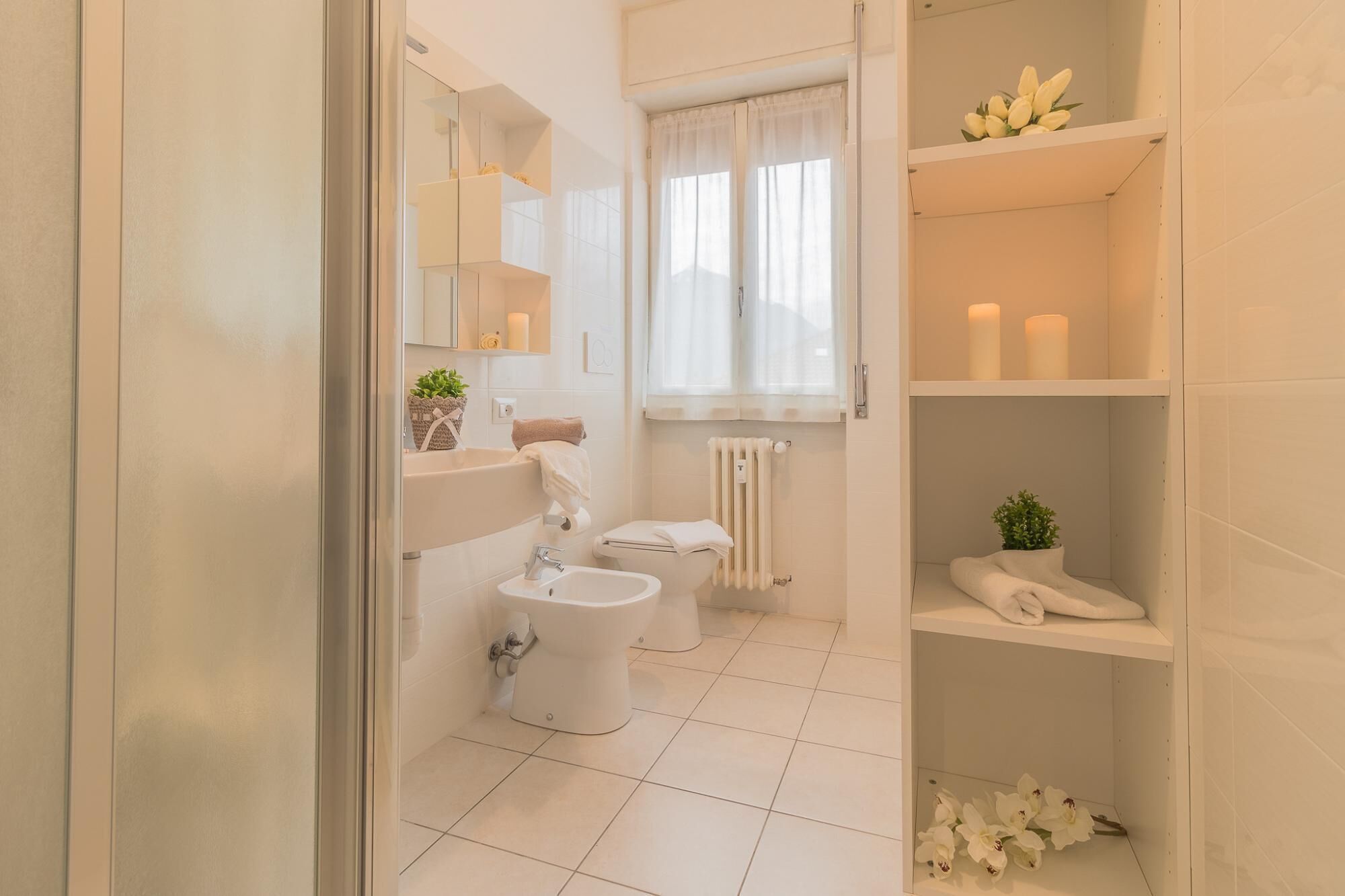 Une salle de bain moderne équipée de toilettes, d’un bidet et d’un lavabo. On y trouve des étagères avec des bougies et une plante.
