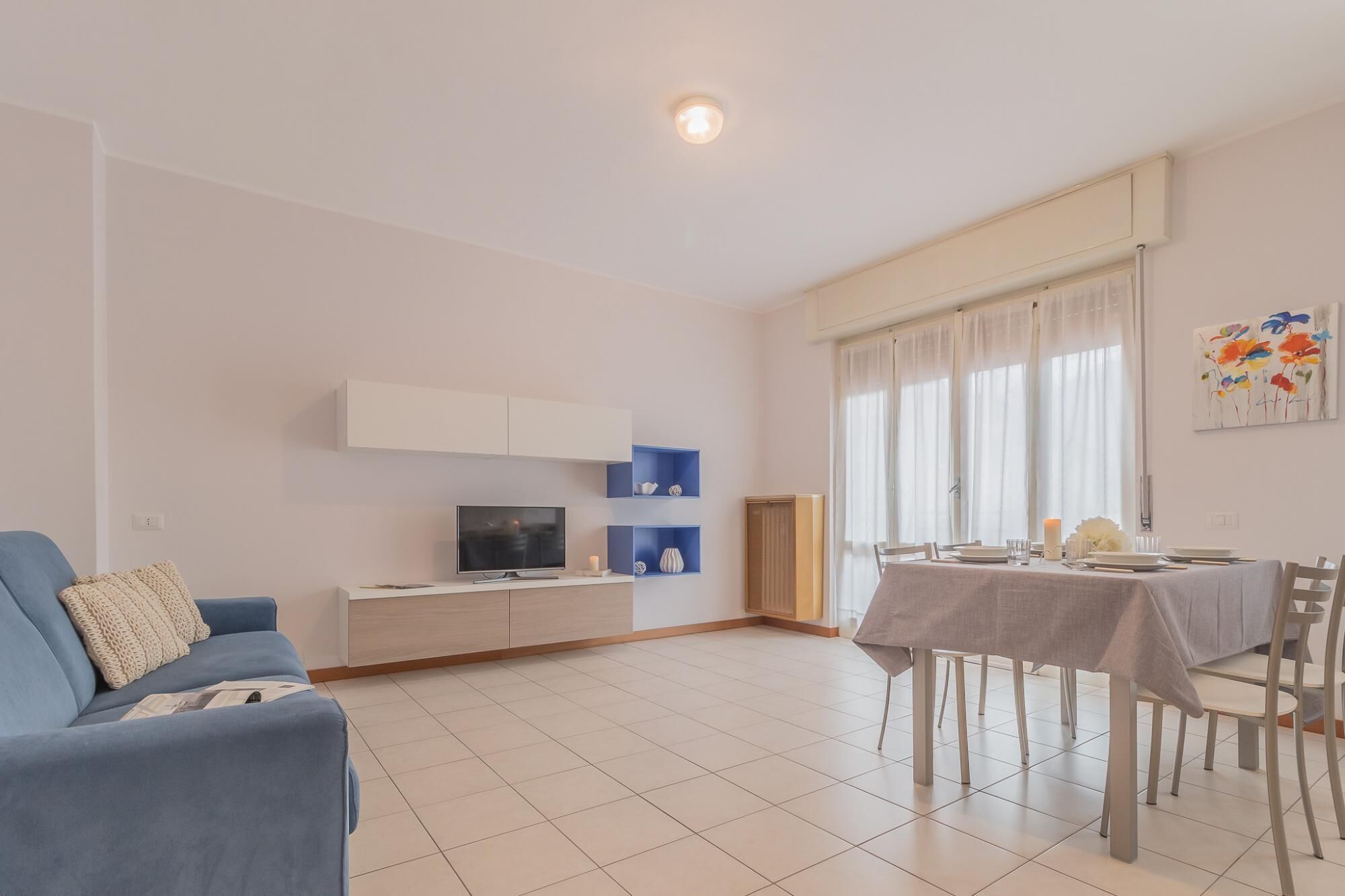A modern living room with a blue sofa, a TV, a dining table set for two, and a large window with curtains.