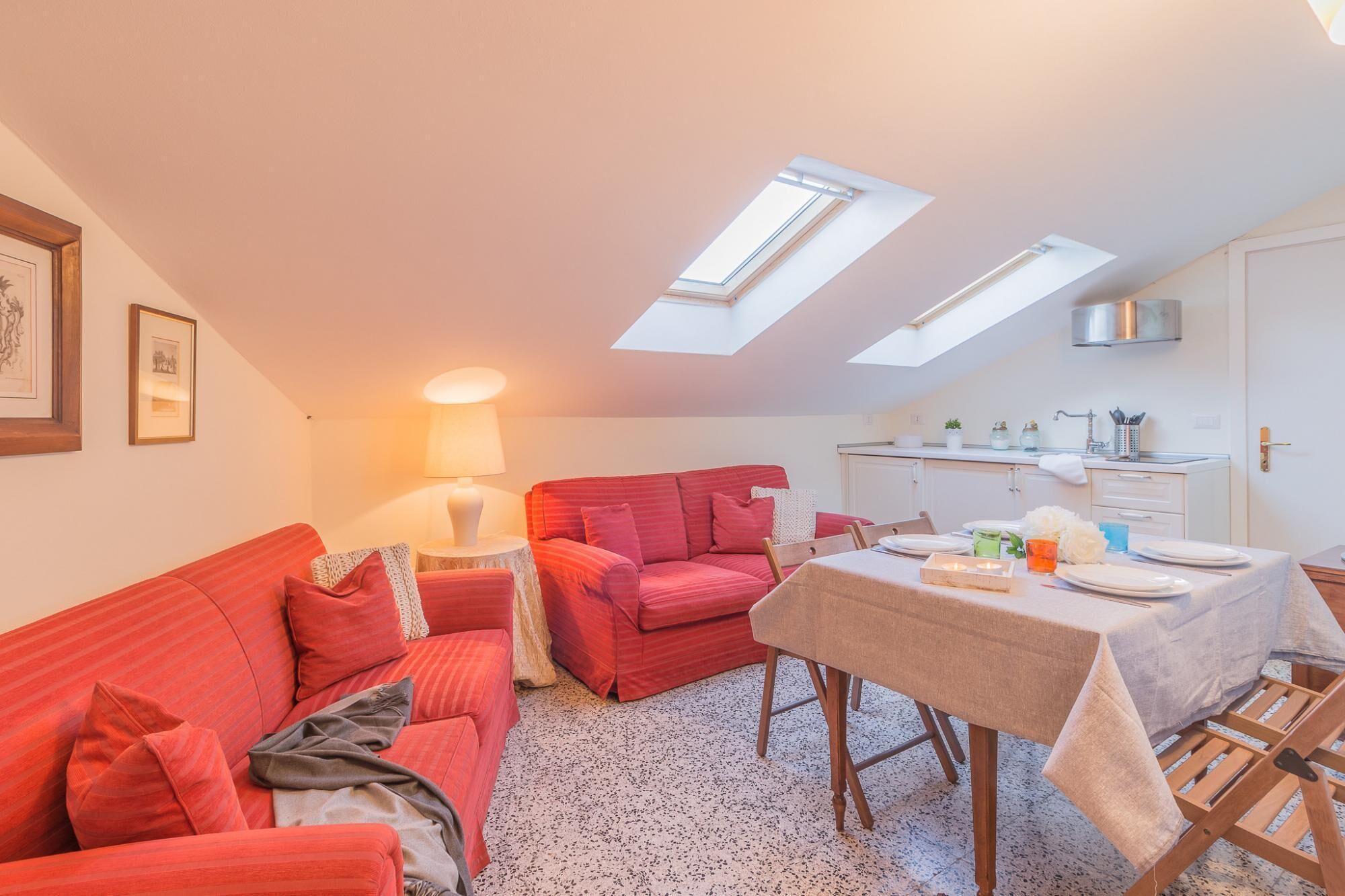 Una sala de estar acogedora con un sofá rojo, una mesa de comedor para dos personas y una cocina con ventana sobre el fregadero.