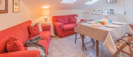 Una sala de estar acogedora con un sofá rojo, una mesa de comedor para dos personas y una cocina con ventana sobre el fregadero.