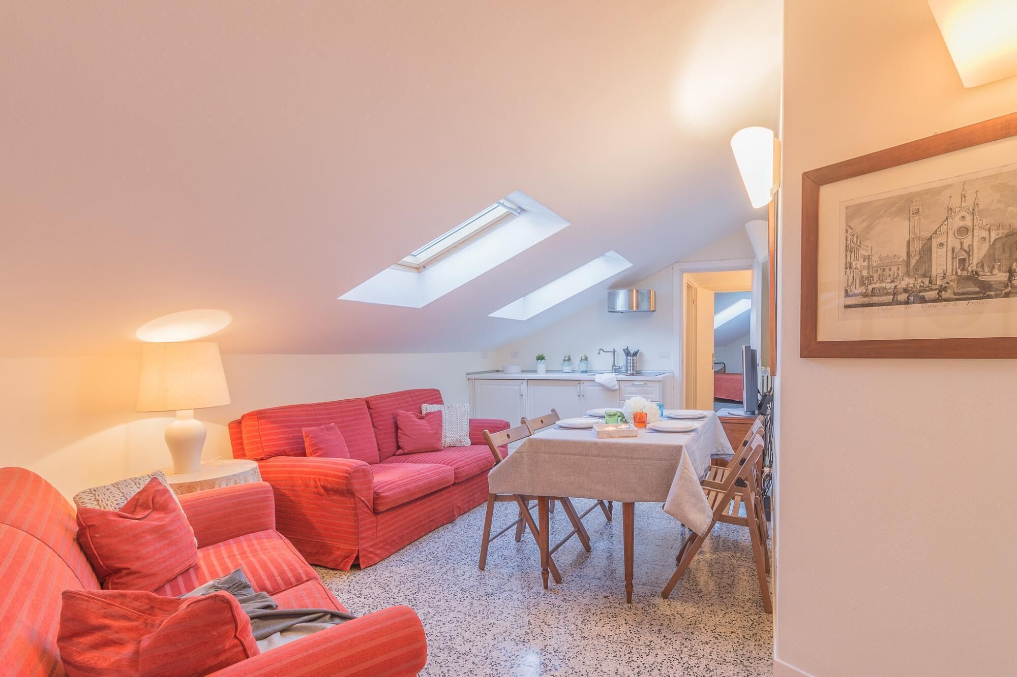 A cozy living room with a red sofa, a dining table set for two, and a kitchen in the background.