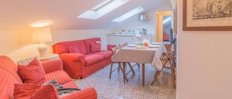 A cozy living room with a red sofa, a dining table set for two, and a kitchen in the background.
