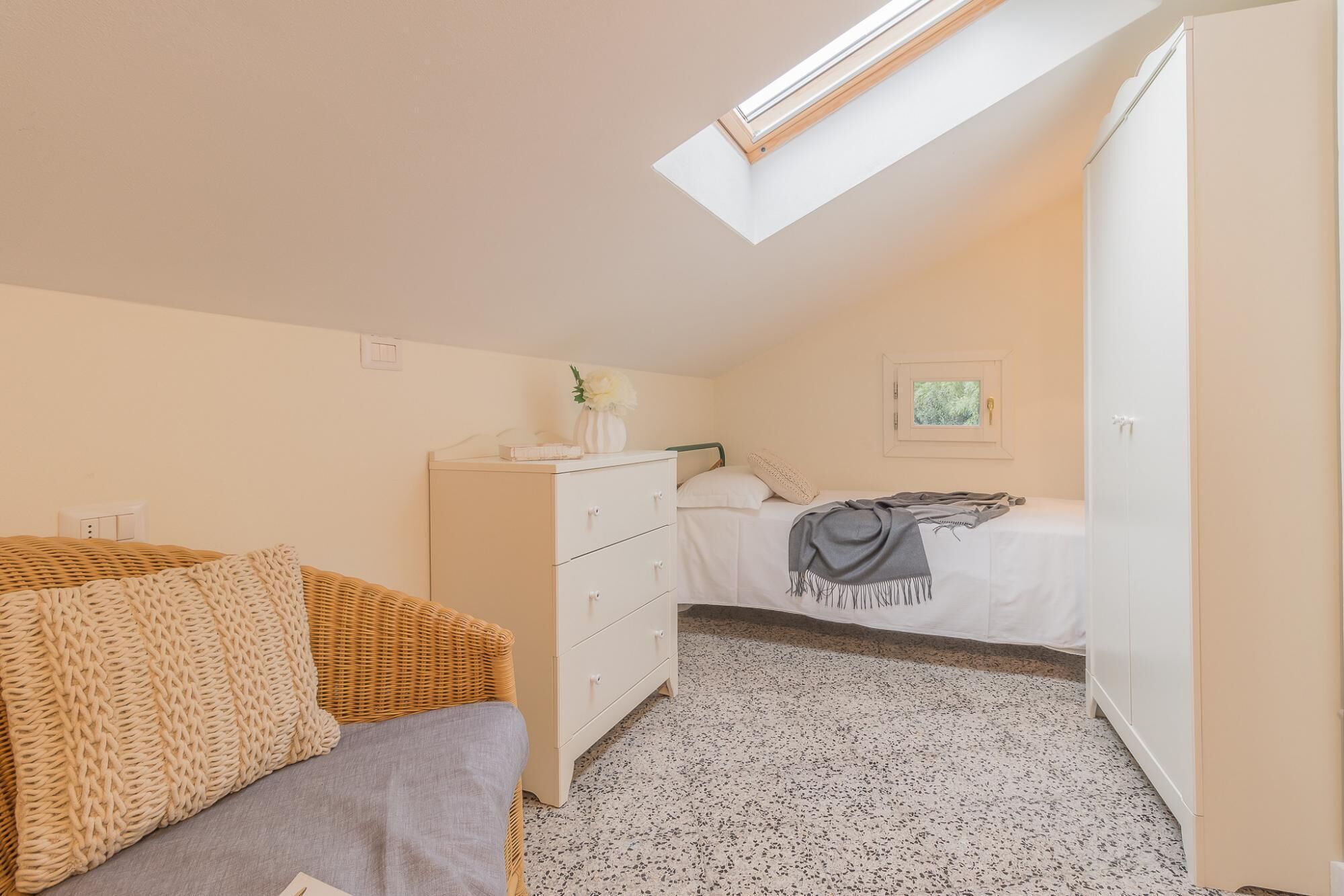 Ein Schlafzimmer mit Dachfenster, einem Bett mit grauer Decke, einem Korbstuhl, einer weißen Kommode und einem kleinen gerahmten Bild an der Wand.