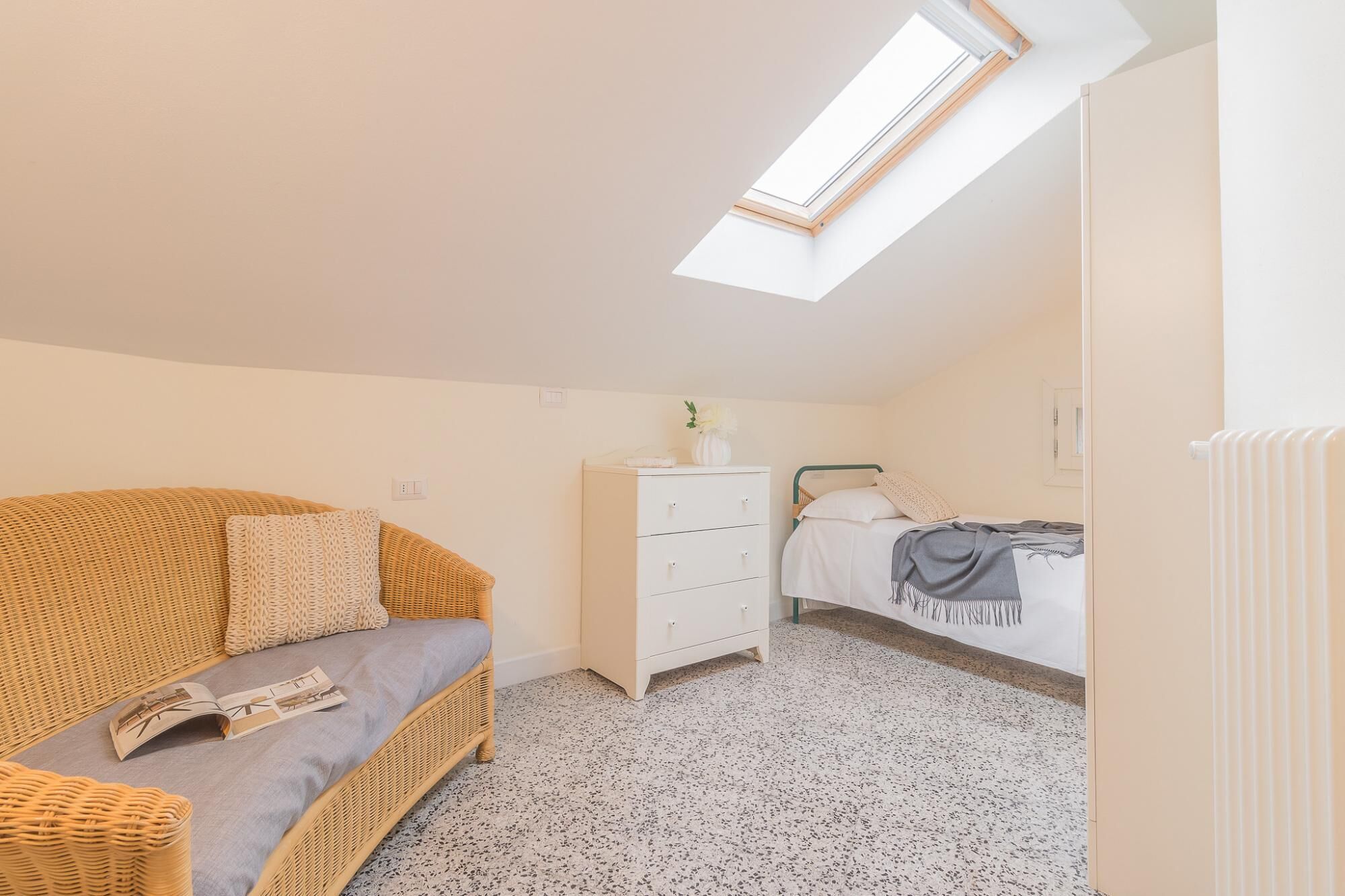 A cozy room with a high ceiling, a skylight, a wicker sofa, a white dresser, and a bed with a grey blanket.