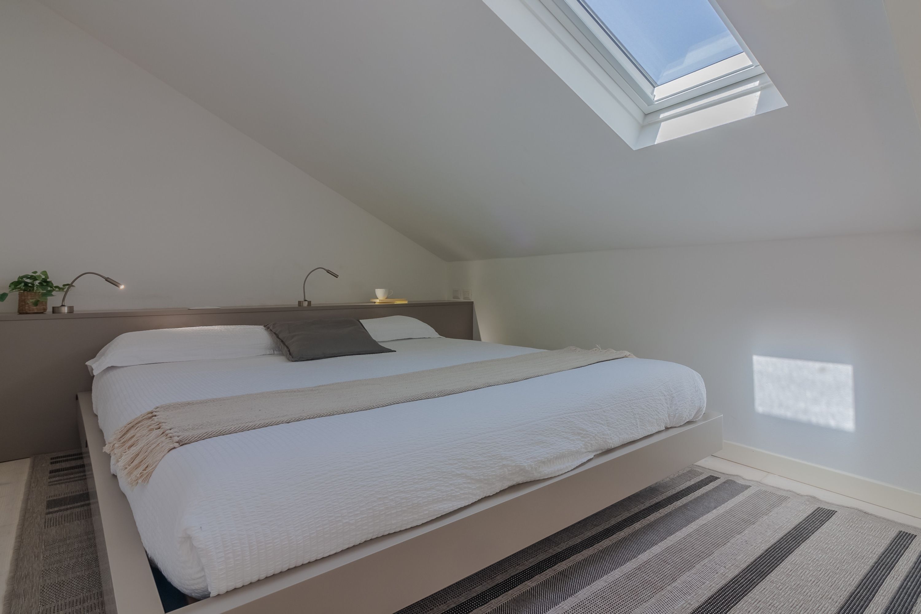 A bedroom with a skylight, a bed with white bedding, a bedside table with a lamp, and a small plant.