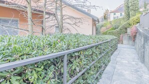 A pathway flanked by a metal railing and dense green foliage, leading to a building with a red-tiled roof and a beige facade.