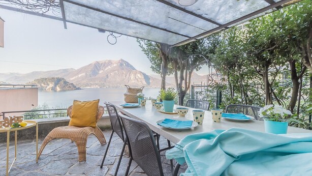 An outdoor patio with a table set for dining, overlooking a body of water and mountains.