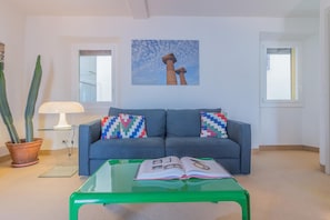 Un salon avec un canapé bleu, deux coussins à motifs, une table basse verte sur laquelle est posé un magazine ouvert, une plante en pot et un tableau accroché au mur.