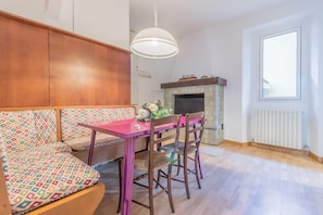 Une pièce chaleureuse avec un banc en bois, une table rose et des chaises. On aperçoit une cheminée au fond, et il y a une fenêtre sur le côté droit.